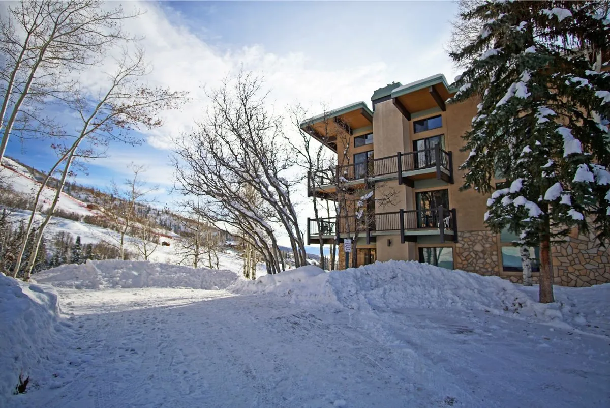 A serene winter exterior scene featuring a snow-covered ground, trees, and the condo building with a cozy balcony overlooking the tranquil snowy landscape. The setting radiates peacefulness and showcases the building's prime proximity to outdoor activities like skiing and snowboarding.