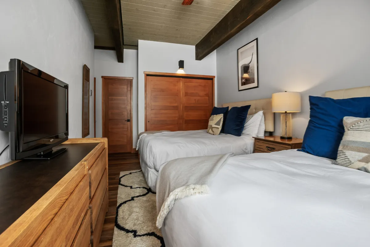 A cozy bedroom with two beds featuring blue accent pillows, wooden furniture, a central lamp, and decorative artwork. The space exudes a sense of comfort and is ideal for friends or family traveling together, offering shared relaxation and privacy.