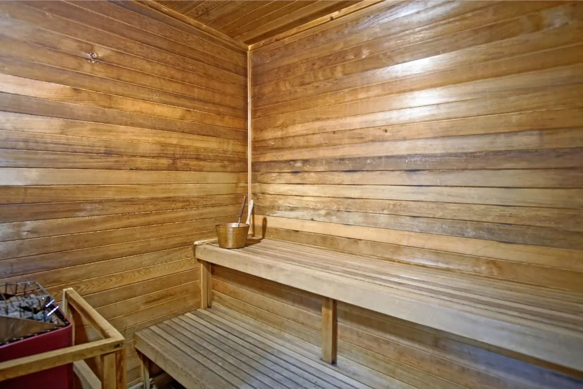 A serene indoor sauna with wooden benches and walls, featuring a classic sauna bucket in the corner. The warm lighting and inviting tone create a peaceful, restorative atmosphere—ideal for relaxation and self-care.