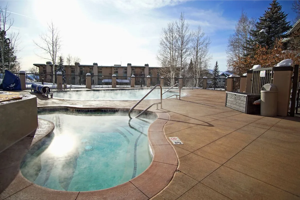 A tranquil hot tub with steam wafting up, surrounded by a wooden fence and tall trees, creating a secluded and rejuvenating space. The warm water contrasts invitingly with the chilly outdoor air, with hints of the sunset peeking through the trees.