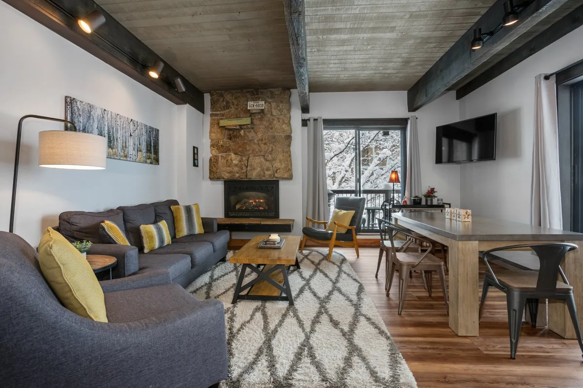 A warm, inviting interior featuring a plush sofa set in front of a glowing fireplace, with a dining table nearby. Large windows frame sweeping mountain views, and natural light floods the space. The setting is perfect for relaxing, socializing, or enjoying meals together while immersed in an alpine atmosphere.