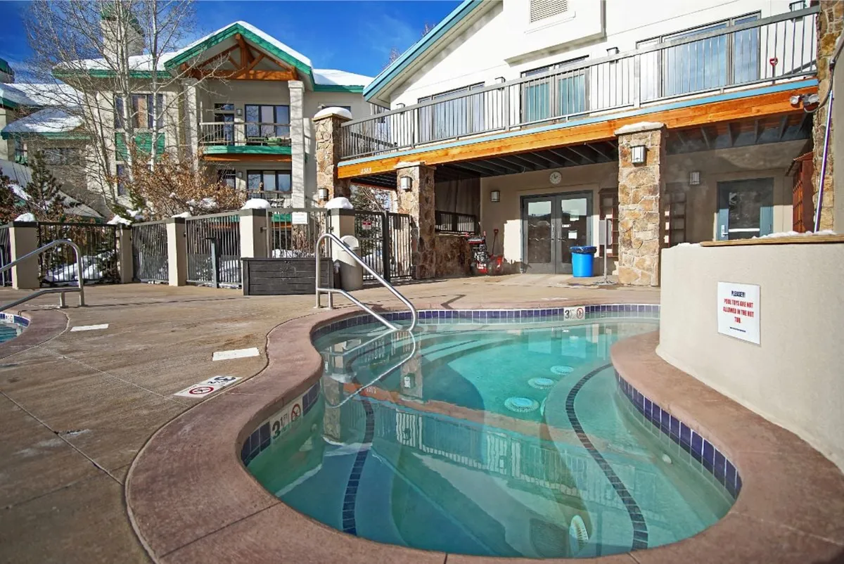 A welcoming exterior shot featuring a hot tub surrounded by snow, the building’s balcony area, and trees. The dusk lighting and inviting hot tub emphasize après-ski relaxation and community gathering after a day on the slopes.