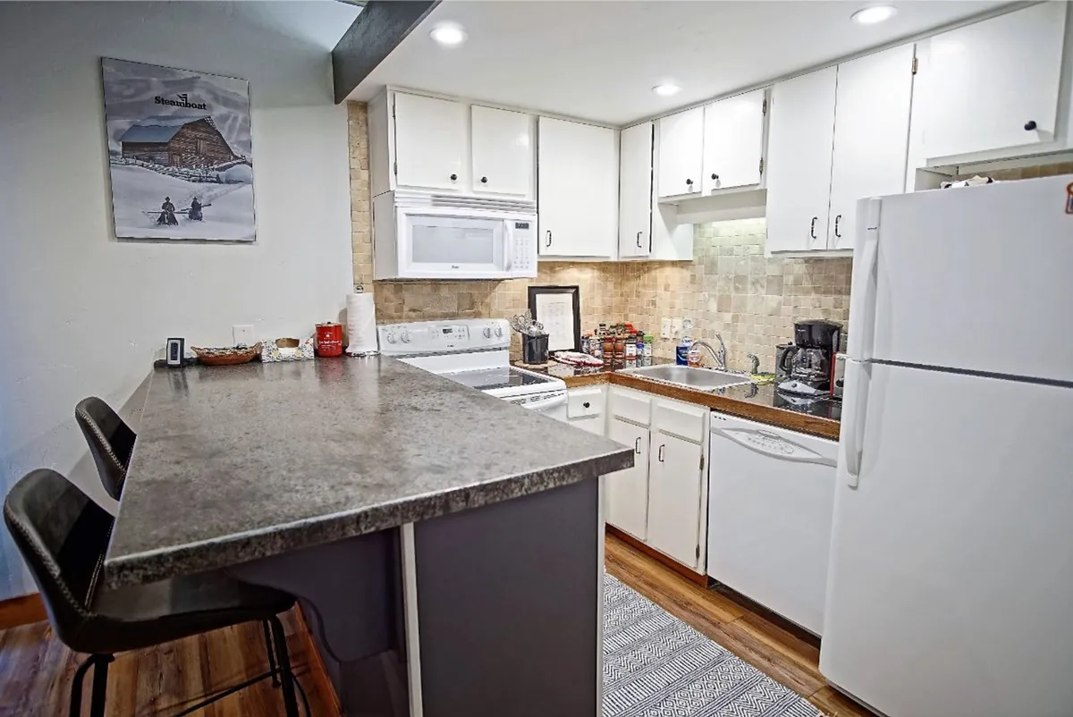 A bright view of the kitchen features a sleek island, crisp white cabinets, modern appliances, generous counterspace, and subtle mountain-inspired artwork. The open layout suggests a welcoming, easy-to-navigate environment for both food prep and gathering.