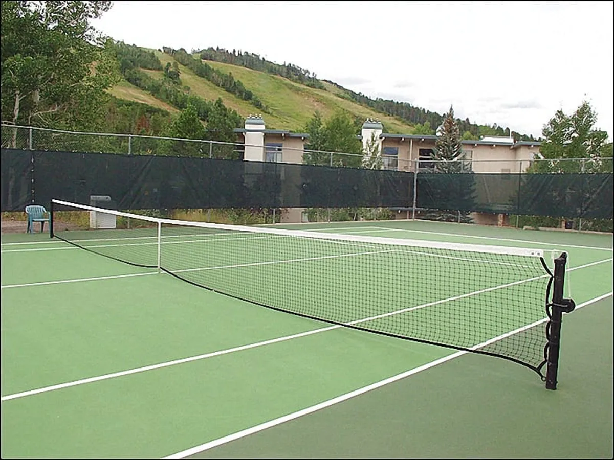 A vibrant outdoor tennis court surrounded by trees and residential buildings, with a clear view of the net and court surface. The atmosphere suggests active, sociable recreation within the development.