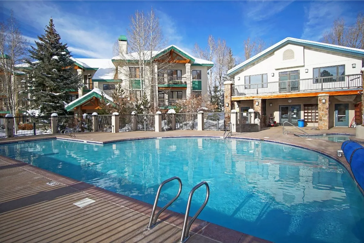 The image showcases the condo’s heated outdoor pool surrounded by lush trees, with the property’s inviting architecture visible in the background. The scene is set at sunset, casting a warm glow over the pool, perfect for swimming, relaxing, or socializing after a day on the slopes.