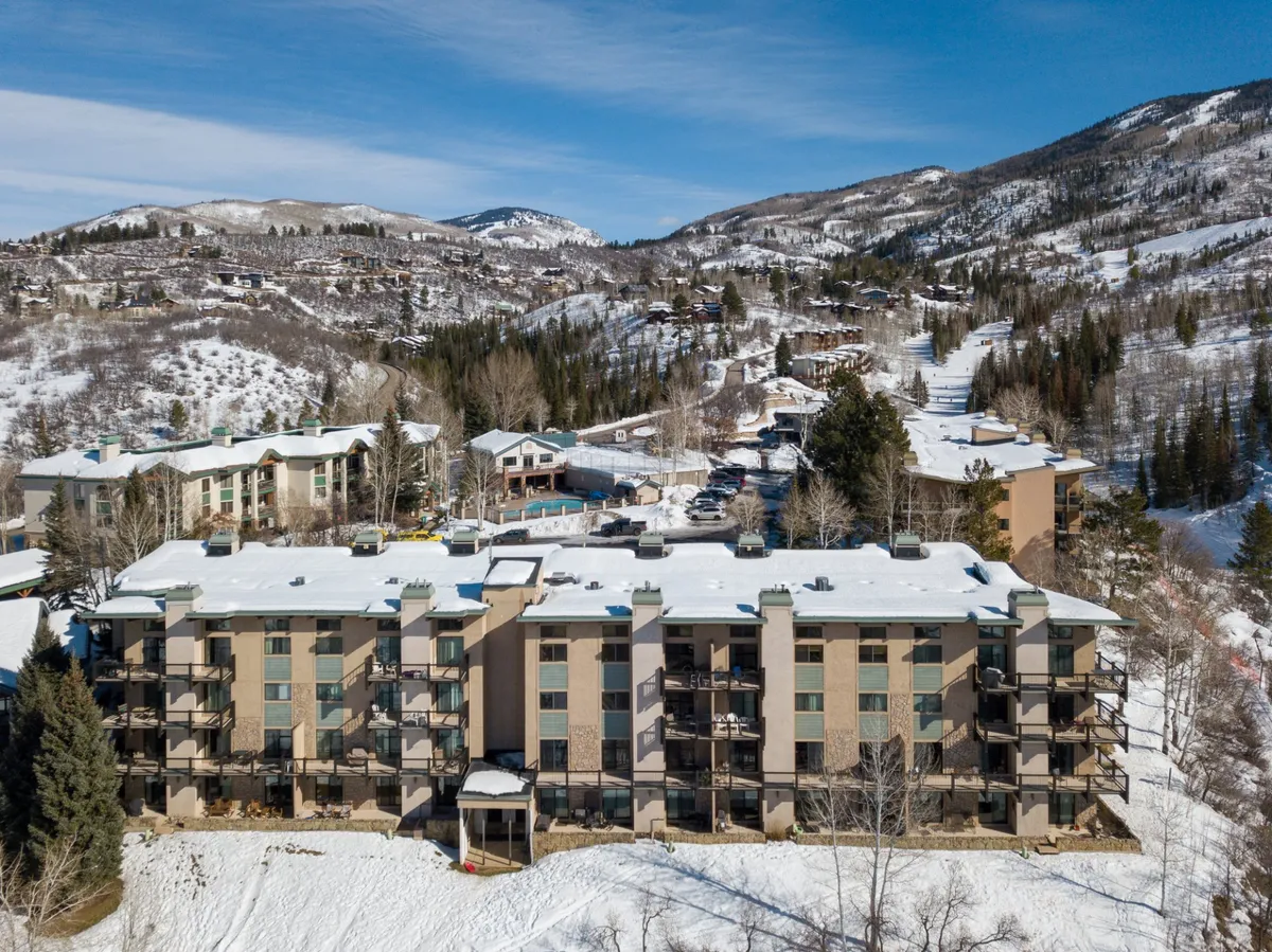 A wide view of a multi-story building set against a backdrop of snow-covered mountains and trees, with visible balconies. The scene suggests a communal, welcoming atmosphere and emphasizes the building's capacity for groups or families.