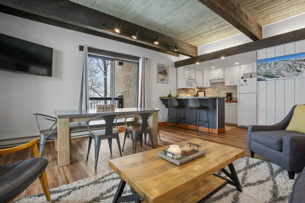 A spacious open-concept living and dining area, featuring a large dining table, kitchen island, and ample seating. Sunlight streams through an expansive window, offering a picturesque mountain view and fostering a sense of togetherness.