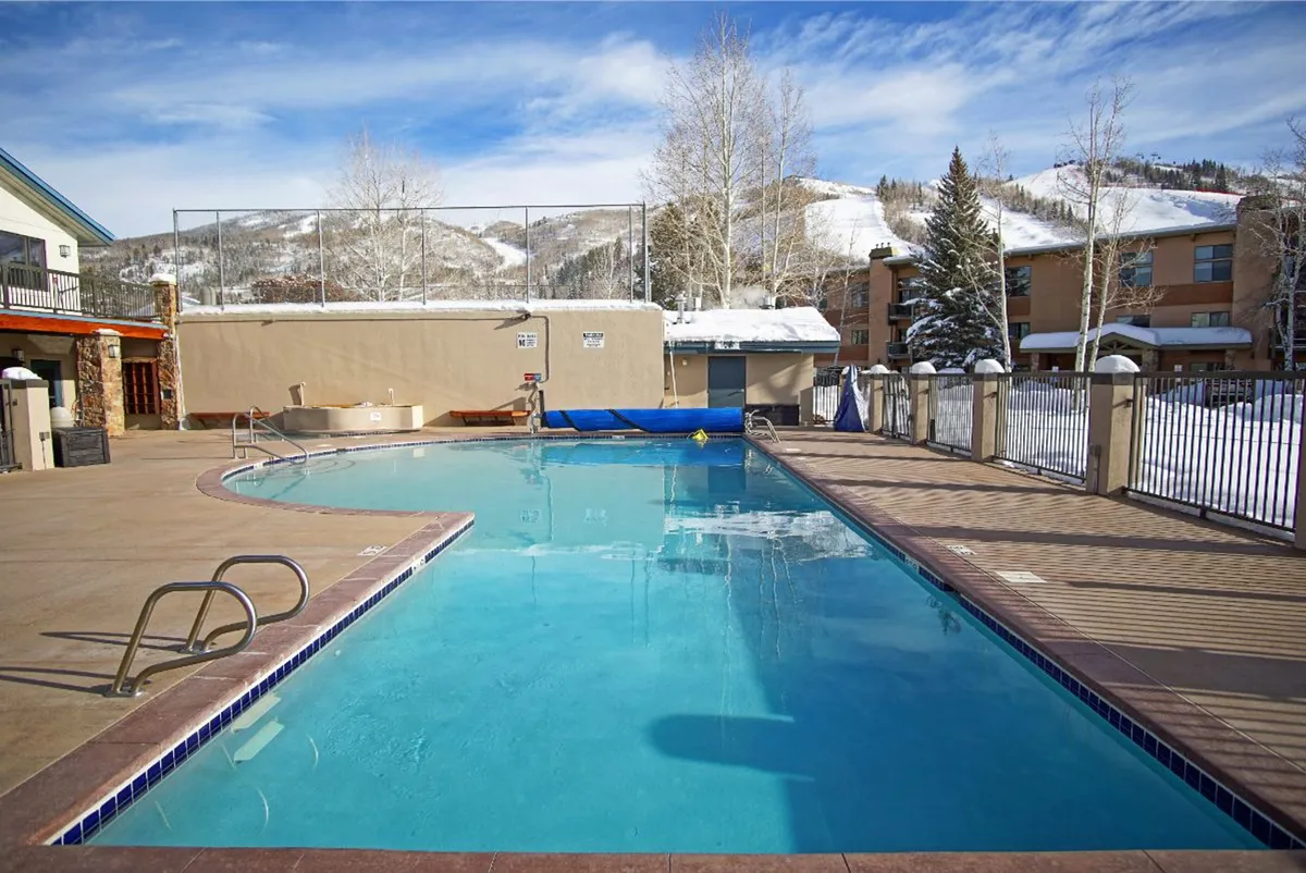 An inviting outdoor heated swimming pool with steam rising from the water, set against a backdrop of snow-covered mountains and tall pine trees. The serene pool area is surrounded by a deck, perfect for lounging after a swim, and offers beautiful sunset and mountain views.