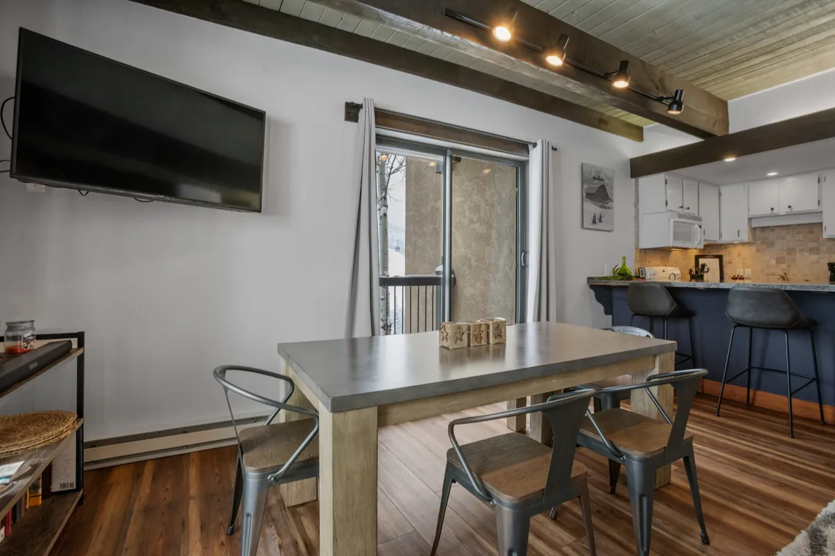 A cozy, sunlit living space with a dining table, TV, and a bar counter. A sliding glass door leads directly to a wraparound deck, hinting at outdoor access and mountain views, with sunlight pouring into the room.