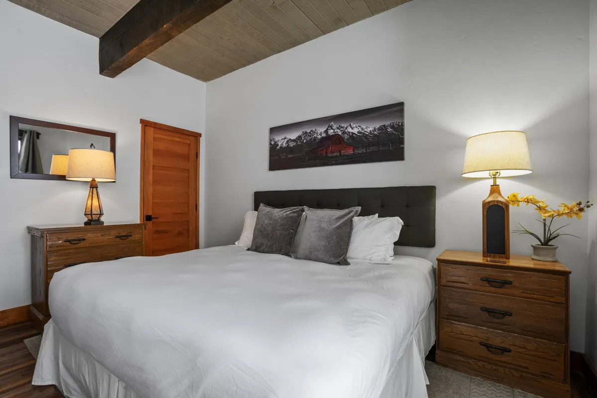 A serene bedroom featuring a spacious king-sized bed dressed in crisp white linens, flanked by wooden nightstands and table lamps. Decorative artwork hangs above the headboard, and natural light filters in, creating a tranquil atmosphere perfect for relaxation and restful sleep.
